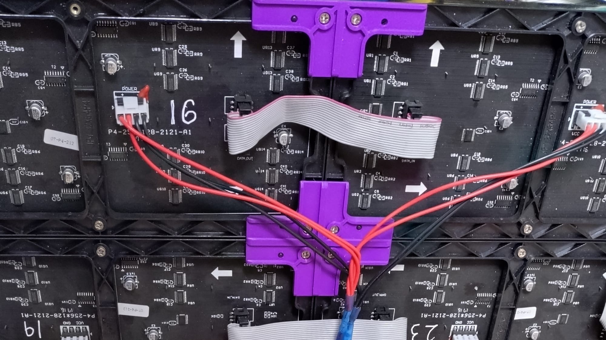 Back view of 4 joined led panels, a grey communications cable joins them, as well as red and black power cables. Importantly there are purple 3d printed brackets holding them together